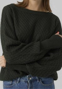Maglione lavorato a maglia verde scuro con un motivo a diamante testurizzato, ampio scollo a barca e polsini a costine. Indossato con jeans in denim blu.