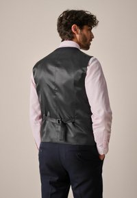 Gilet en satin noir à la texture lisse, avec une ceinture réglable au dos, porté sur une chemise à carreaux rose clair, associé à un pantalon bleu marine.