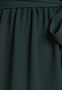 Ciemnozielony, teksturowany materiał z marszczonym wzorem, z szerokim paskiem w talii i związanym detalem. Gładka powierzchnia z miękkim drapowaniem.