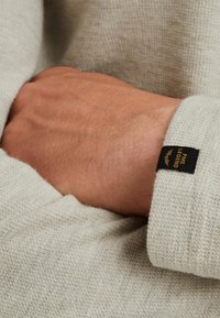 Light gray textured fabric sweater featuring a visible black label on the cuff with "Pine Legend" text. Hand resting against the sleeve.