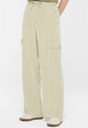 Beige weite Cargo-Hosen mit Knöpfen an den seitlichen Taschen, getragen mit weißen Sneakern, an einer Person stehend vor einem weißen Hintergrund.