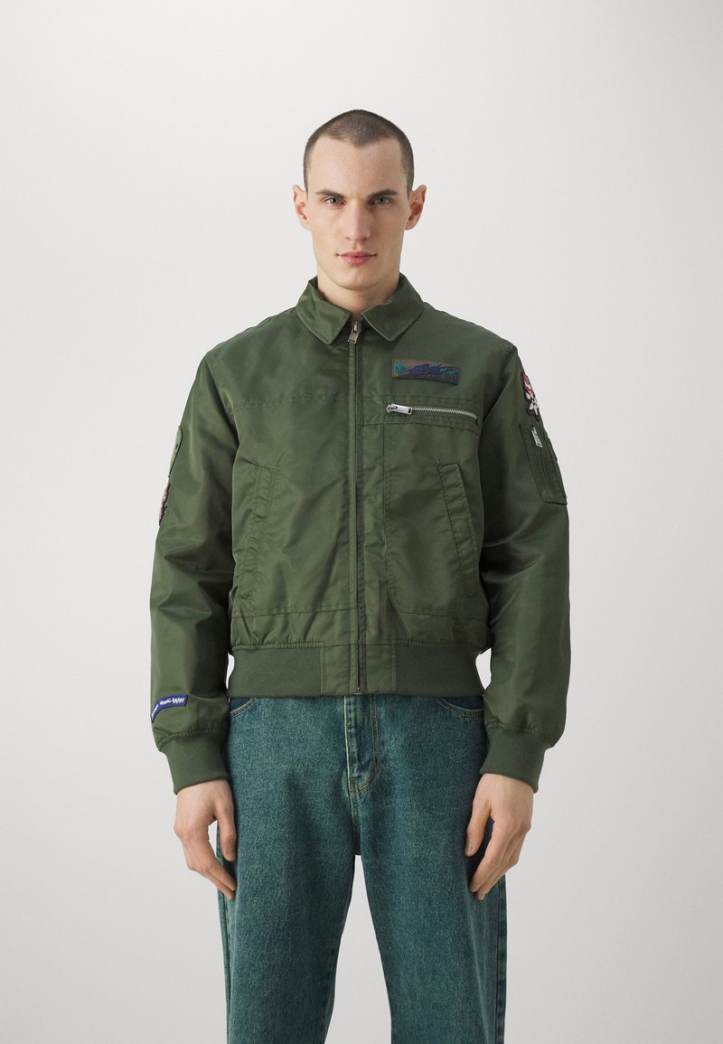 Giacca bomber verde oliva realizzata in materiale leggero, con tasca con zip, toppe ricamate e orlo a costine.