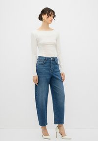 Top blanco de manga larga y jeans de mezclilla azul de corte alto con un ajuste relajado, que cuentan con bolsillos frontales y cierre de botón. Elegantes tacones blancos.