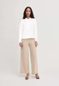 Blouse blanche à manches longues avec un détail de nœud papillon, assortie à un pantalon large beige. Les textures lisses et les couleurs neutres dominent l'ensemble.