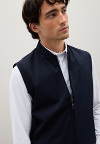 Navy ärmellose Weste aus glattem Stoff mit Reißverschluss, hohem Kragen und betonten Armausschnitten, darüber getragen über einem weißen Langarmshirt.