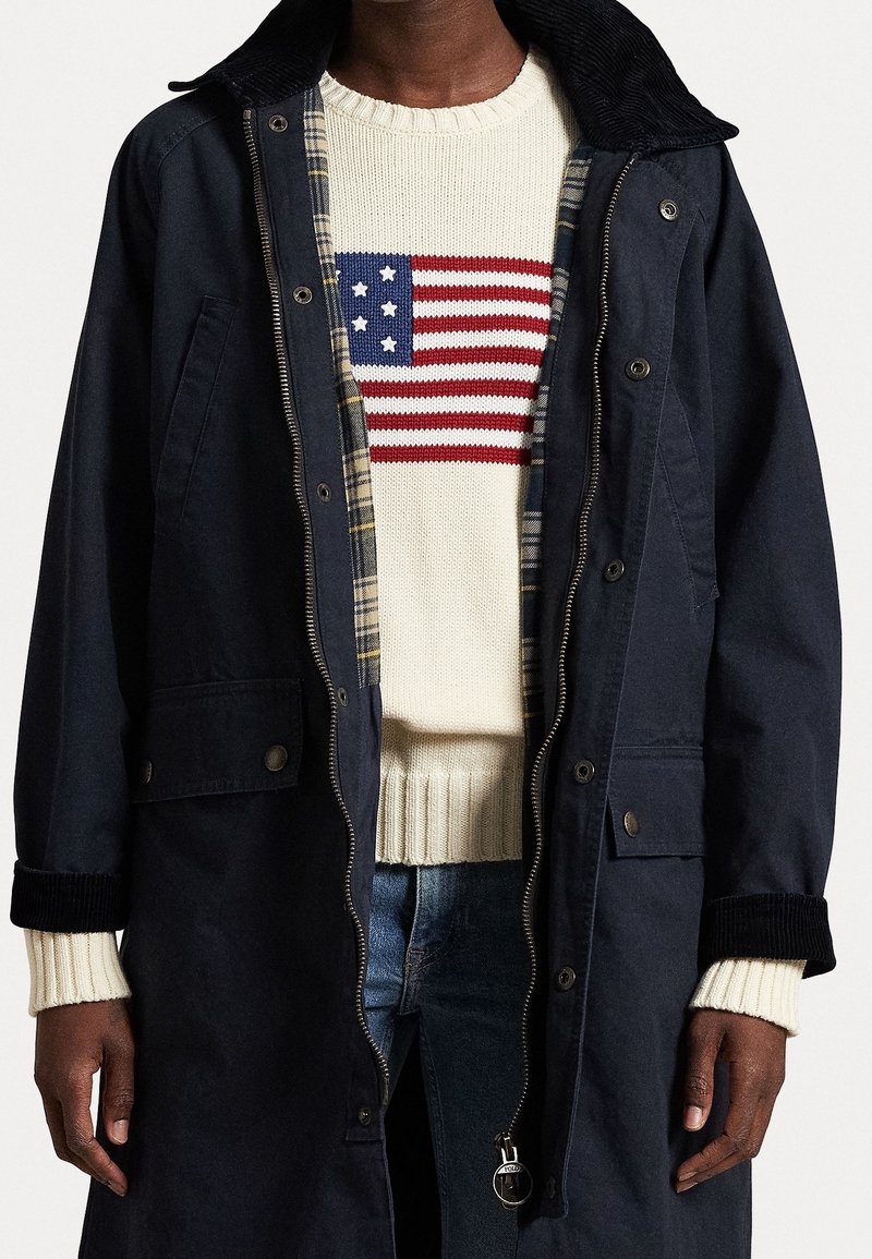 Chaqueta exterior de navy con cuello de pana, cierre de cremallera y bolsillos, superpuesta a un suéter crema con un diseño de la bandera estadounidense en rojo y azul.