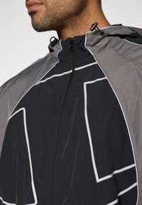 Chaqueta ligera con capucha en negro con acentos grises, que presenta un cierre frontal, líneas blancas en contraste y un acabado de tejido texturizado.