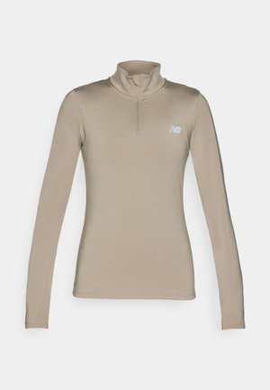 Haut de sport à manches longues beige avec un col montant et une fermeture éclair quart. Il présente un petit logo bleu clair sur le côté gauche de la poitrine. Texture lisse.