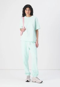 Muntgroene katoenen t-shirt en bijpassende joggerbroek, relaxte pasvorm. Voorzien van een elastische tailleband met trekkoord. Gecombineerd met lichtgrijze sneakers en een roze tas.