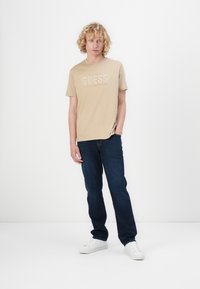 Beige bomulls t-skjorte med "GUESS" logo i hvitt, kombinert med mørkeblå denimjeans og hvite joggesko. Enkelt design, avslappet passform.