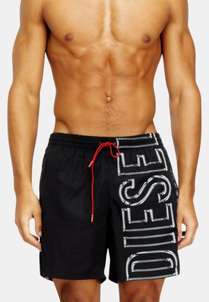 Skjorteløs mand iført sorte badeshorts med stor hvid "DIESEL" tekst og en rød snøre på en enkel lys baggrund.