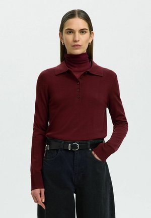 Femme aux cheveux raides portant un polo bordeaux à manches longues, un jean noir et une ceinture noire, debout avec une main dans la poche.