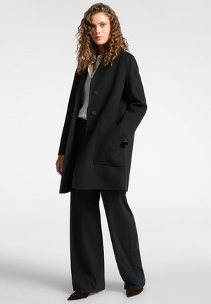 Cappotto di lana nero con taglio dritto, tre bottoni e una tasca. Indossato sopra una camicia a righe, abbinato a pantaloni neri e scarpe scure.