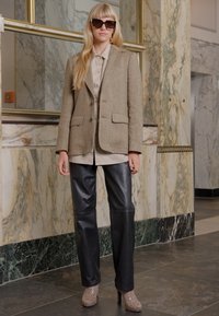 Blazer en tweed beige avec deux poches avant, associé à un pantalon en cuir noir. Le mannequin porte des lunettes de soleil oversize et des escarpins beiges.