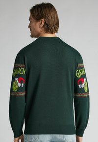 Pull en maille verte avec un motif du Grinch sur les manches, orné de motifs rouges et verts. Ourlet et poignets côtelés.