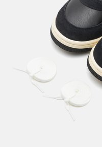 Sorte ruskind- og lædersneakers med cremede gummisåler, parret med to hvide runde snørebånd. Nærbillede viser tekstur og detaljer.