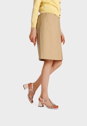 Donna che indossa una gonna beige a lunghezza ginocchio, un maglione giallo e sandali con tacco basso a blocco, punta aperta e cinturini alla caviglia.