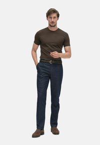 Homme debout avec la main gauche dans la poche, portant une chemise ajustée à manches courtes marron, un pantalon bleu, une ceinture tressée et des chaussures en suédine marron.