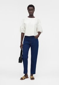 Femme debout, une main dans la poche, portant un haut blanc à manches bouffantes, un jean bleu foncé, des sandales marron et tenant un sac à main noir.