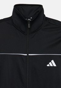 Veste de sport noire avec un col haut, présentant une fermeture éclair sur le devant, une bande horizontale blanche et un logo Adidas blanc en bas à droite.