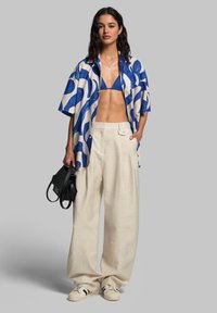 Blue and white patterned short-sleeve shirt, navy bikini top, beige wide-leg pants, holding a black bag, wearing beige sneakers.