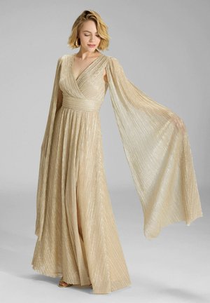 Žena v trblietavých zlatých plisovaných šatách so širokými, plynulo padajúcimi rukávmi, stojaca na jednoliatom sivom pozadí.