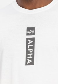 Hvid bomulds t-shirt med et vertikalt sort grafik. Ordet "ALPHA" er tydeligt vist i fed, moderne skrifttype.