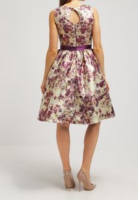 Nainen, joka pukeutunut hihattomaan, kukkakuvioiseen polvipituiseen mekkoon, jossa avonainen selkäosa ja violetti vyötärönauha, yhdistettynä beigen sävyisiin korkokenkiin.