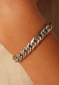 Zilveren kettingarmband met onderling verbonden schakels met een gladde afwerking, gedragen om een gebruinde pols, tegen een gestructureerde lichte achtergrond.
