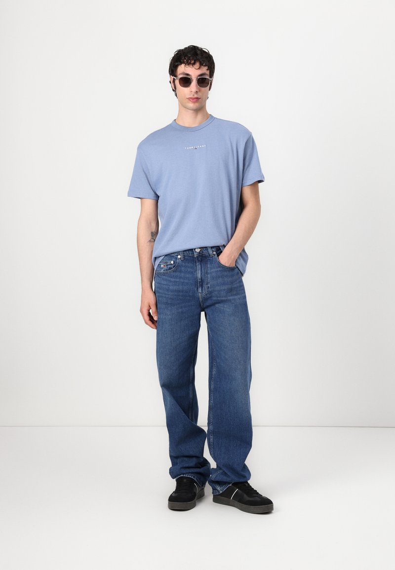 T-shirt en coton bleu clair avec un col rond, assorti à un jean bleu à jambe droite. La tenue est complétée par des baskets noires et des lunettes de soleil.