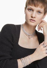 Ein Model mit kurzen Haaren trägt mehrere funkelnde silberne Halsketten und Armreifen, die eleganten Schmuck vor einem schlichten weißen Hintergrund präsentieren.