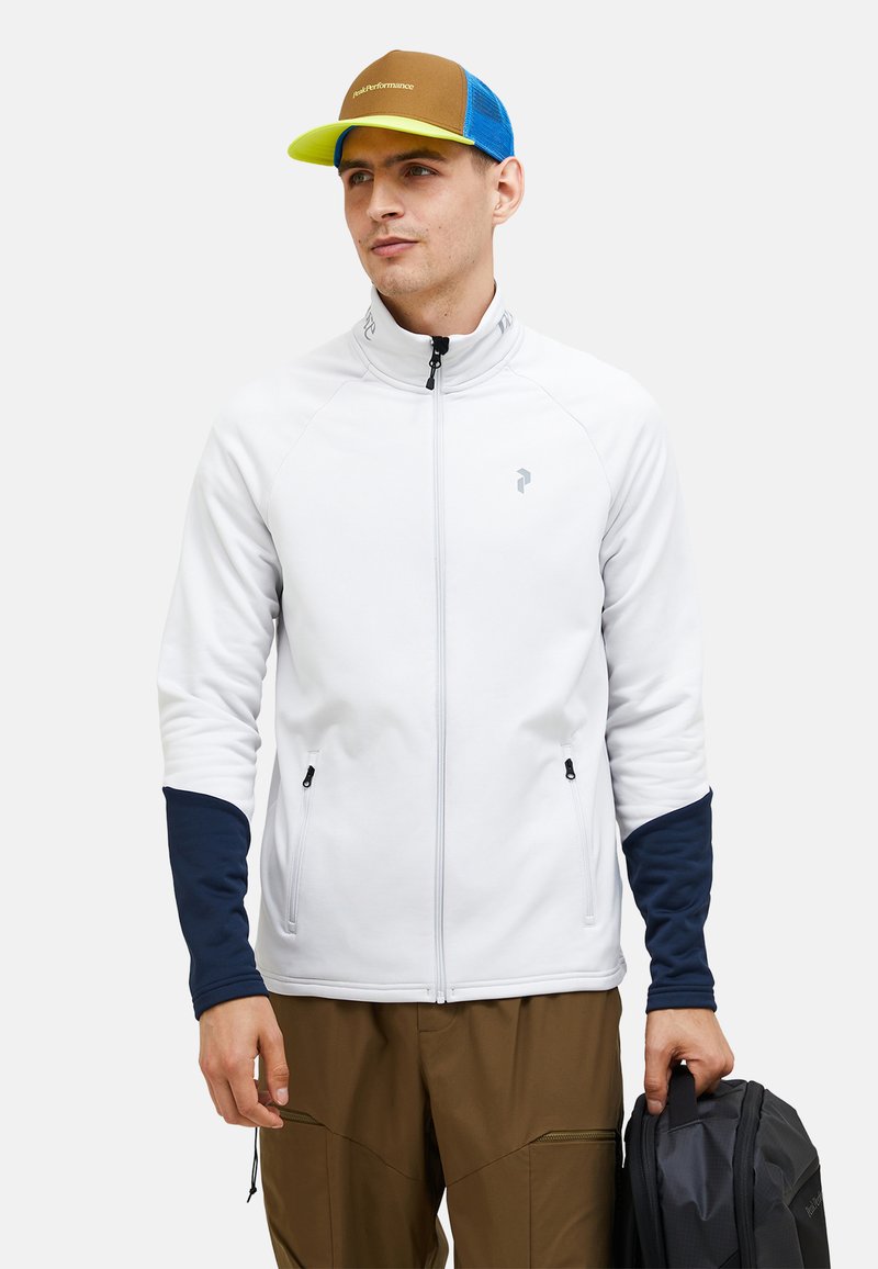 Hvid zip-up jakke med marineblå ærmer, med to sidelommer og høj krave. Personen holder en mørk taske og har en multicolor kasket på.