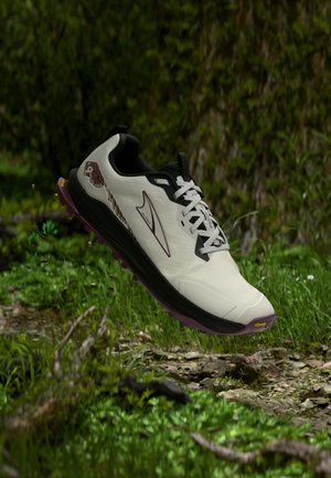 Scarpa da trail running bianca e nera con suola viola che fluttua sopra un sentiero roccioso e erboso all'aperto circondato da fogliame verde.