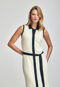 Vestido de longitud hasta la rodilla en crema con canalé vertical, botones negros y un cuello y cinturón en contraste negro. Textura suave, diseño sin mangas.