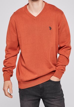 Mann trägt einen rostorangefarbenen V-Ausschnitt-Pullover mit kleinem schwarzem gesticktem Logo auf der linken Brust, kombiniert mit dunkelgrauen Hosen.