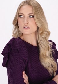 Paarse gebreide trui met ribbeltextuur en gerimpelde schouderaccenten. Glad, golvend blond haar valt voorbij de schouders. Lichte achtergrond.