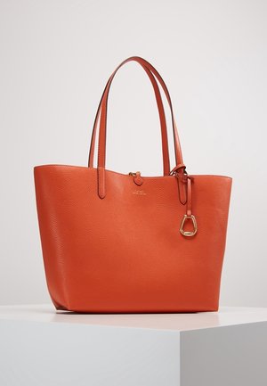 Pomarańczowa torba na ramię z teksturowanej skóry z podwójnymi rączkami i małym złotym ozdobnym charmem, prezentowana na białym podeście na jednolitym tle.