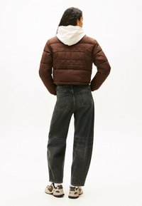 Giacca imbottita marrone con design corto, abbinata a jeans in denim scuri e sneakers beige, con un cappuccio bianco che sbircia.