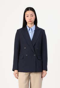 Blazer doppiopetto blu navy con revers a punta, due tasche frontali e bottoni neri, abbinato a una camicia azzurra con colletto e pantaloni beige.