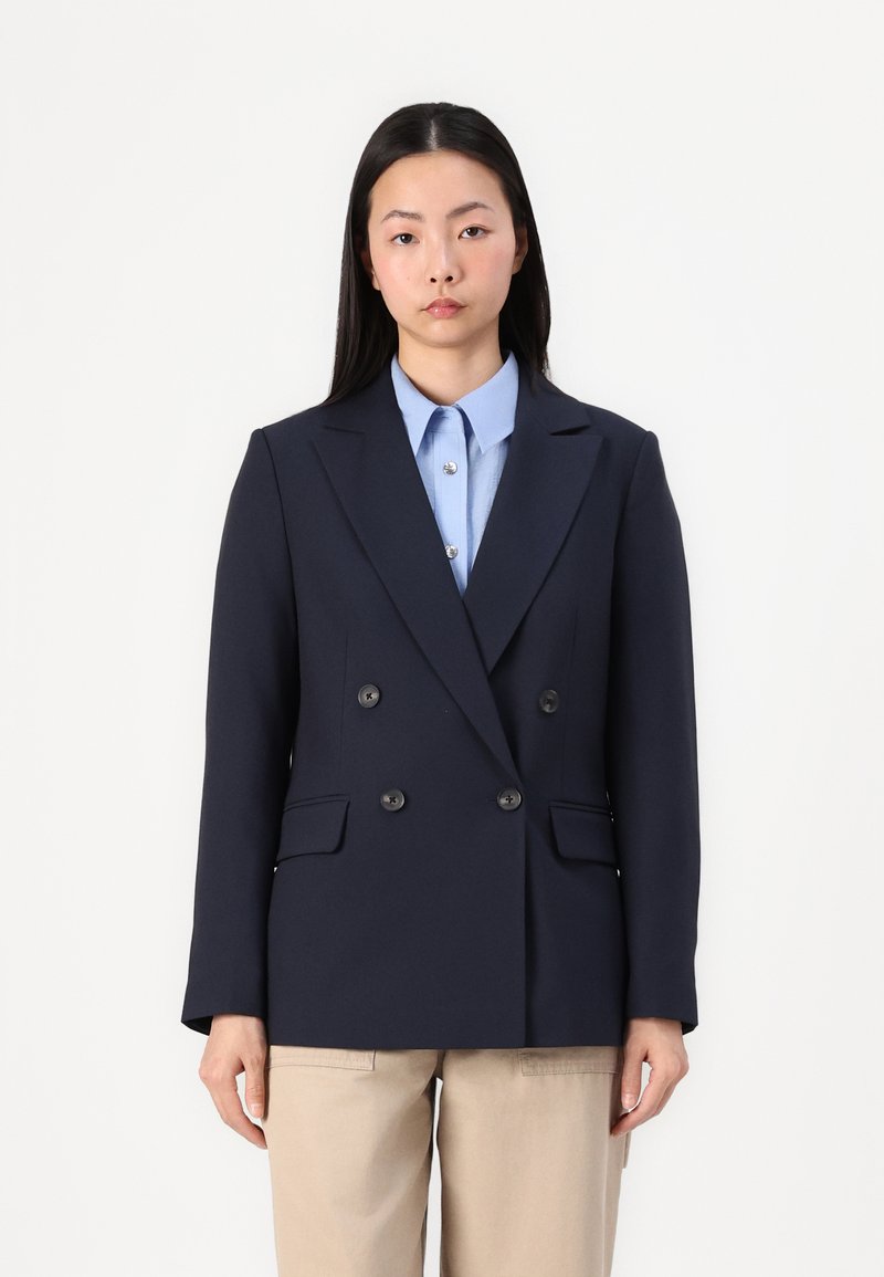 Blazer doppiopetto blu navy con revers a punta, due tasche frontali e bottoni neri, abbinato a una camicia azzurra con colletto e pantaloni beige.