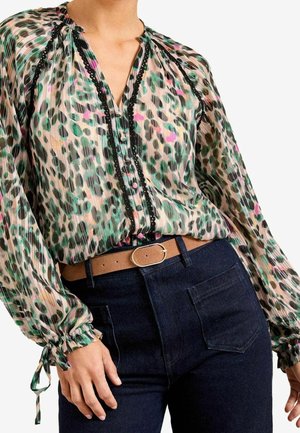 Kvinde iført en gennemsigtig bluse med leopardprint med sort kant og knapdetaljer, kombineret med mørke jeans og et tan-farvet bælte med guldlås.