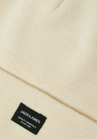 Berretto a maglia color crema con texture a coste e orlo piegato. Presenta un'etichetta nera con testo bianco: "JACK & JONES QUALITY ESSENTIALS SINCE 1990."