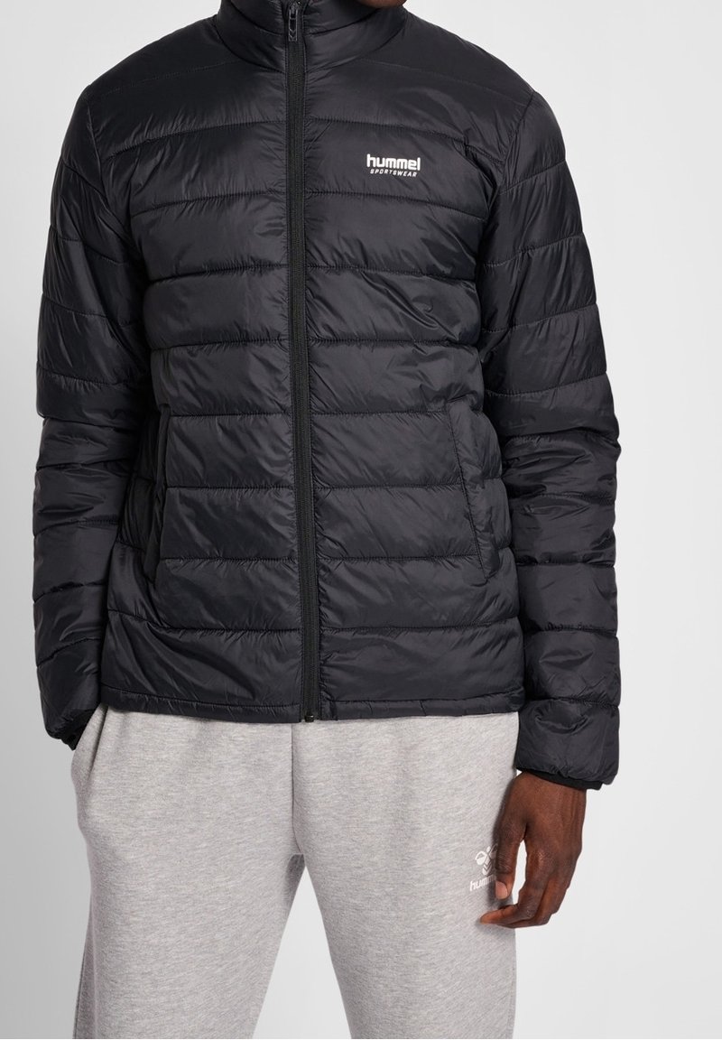 Veste doudoune noire avec un design matelassé, col haut et fermeture éclair intégrale. Présente un petit logo blanc sur la poitrine gauche. Fabriquée en tissu brillant.