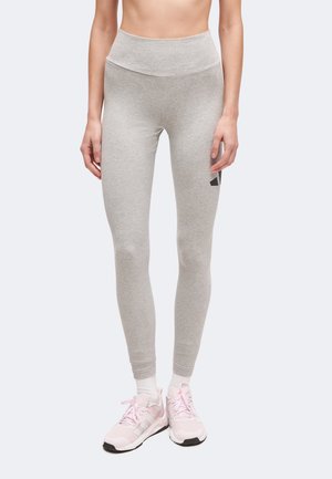 Kvinde iført lysegrå højtaljede leggings og lyserøde sportssko står mod en ensfarvet baggrund.