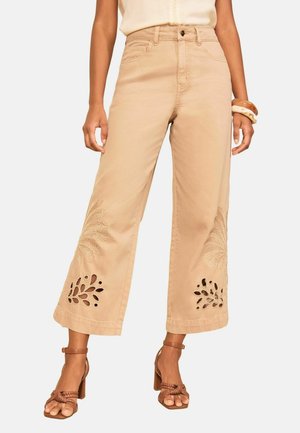 Kvinde iført beige cropped vidde bukser med blomsterudskæringer og broderidetaljer nær kanterne, kombineret med brune sandaletter med hæl.