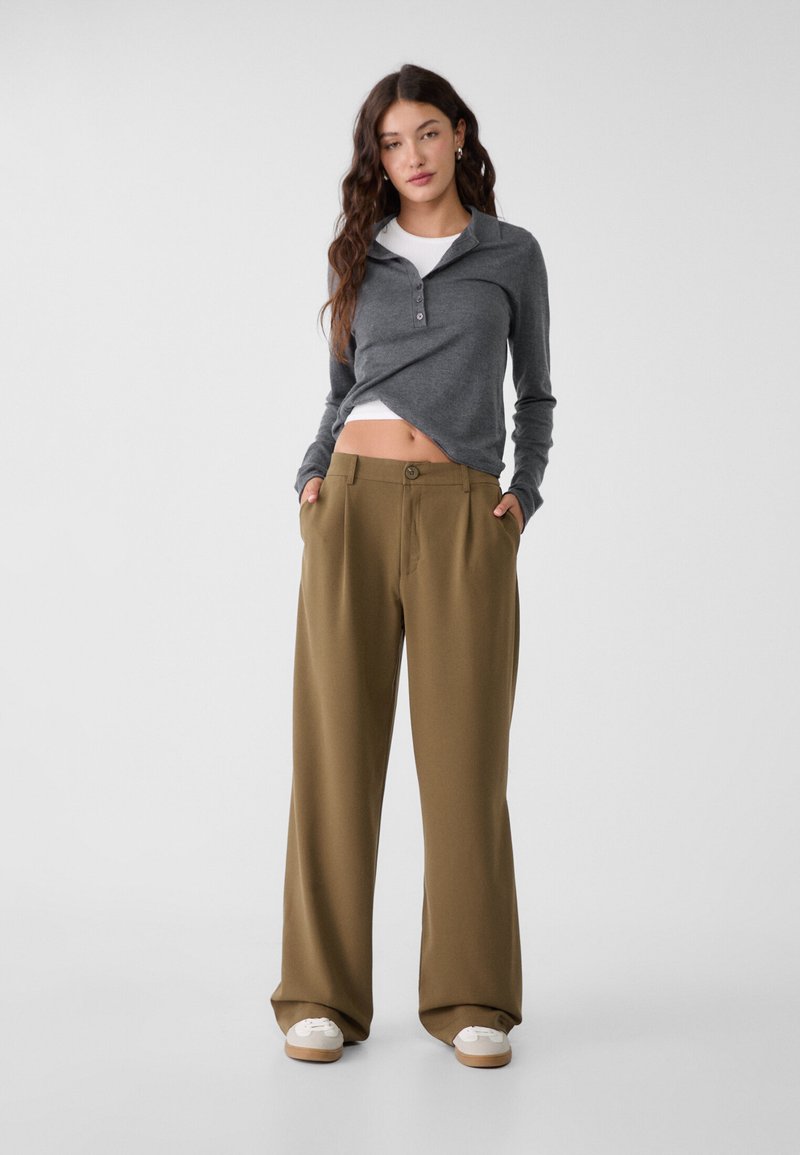 Chemise grise à manches longues avec patte de boutonnage, superposée sur un haut blanc. Pantalons amples beige avec poches latérales. Chaussures blanches avec semelles brunes.