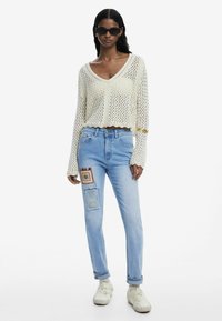 Haut en crochet blanc avec décolleté en V et ourlet à volants ; jeans en denim bleu clair avec des détails de patchwork et de broderie ; baskets blanches.