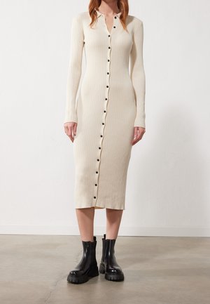 Robe midi en maille côtelée crème avec manches longues, col, et détails de boutons noirs sur le devant, associée à des bottines noires épaisses.