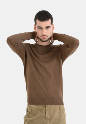 Giovane uomo con capelli corti e scuri, indossa un maglione marrone a maniche lunghe e pantaloni beige, posando con le mani dietro il collo su uno sfondo semplice.