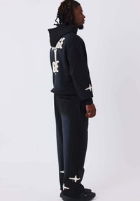 Sudadera negra con texto blanco en la parte posterior y las mangas, combinada con pantalones negros que presentan patrones abstractos blancos; material casual, ajuste relajado.